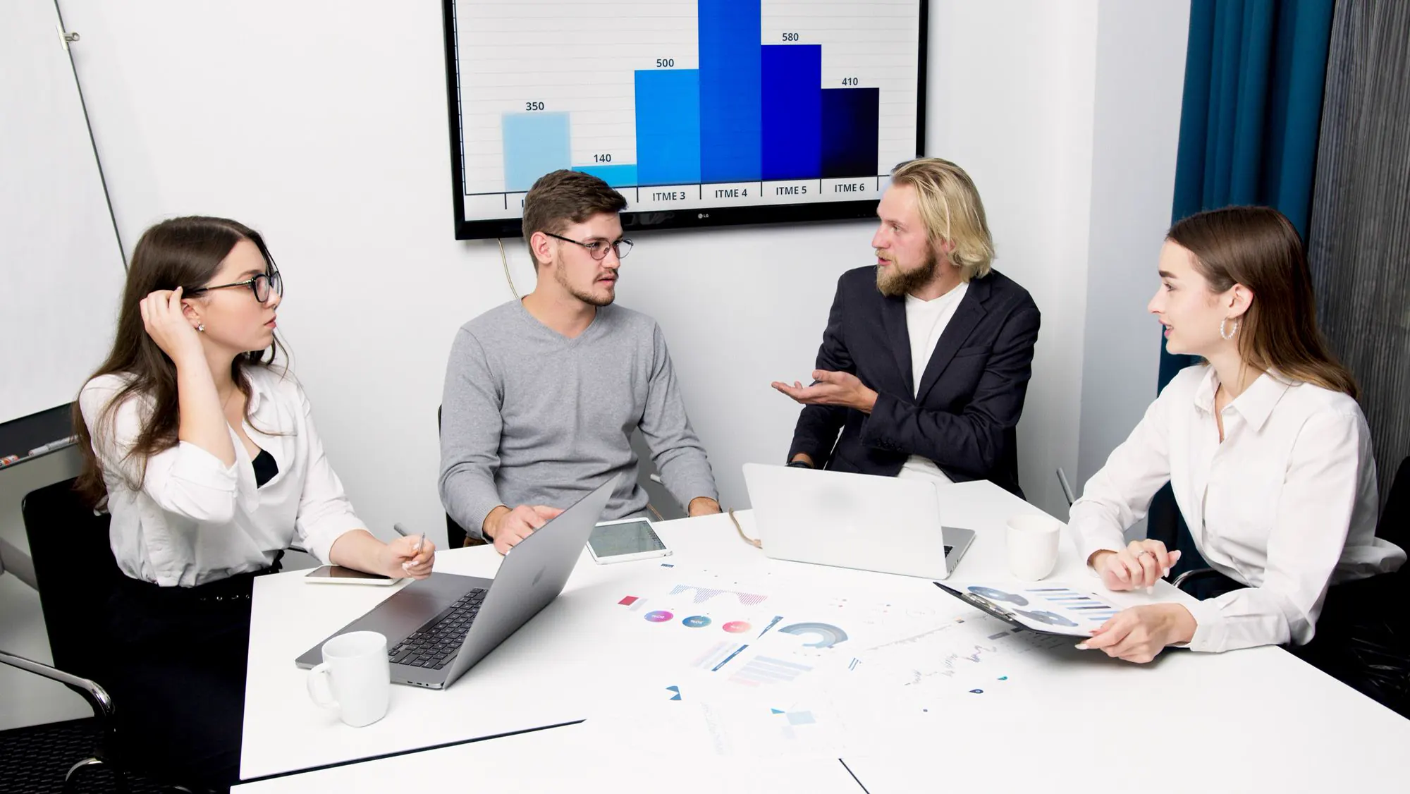 Business team discussing operating results around a meeting table with charts on screen
